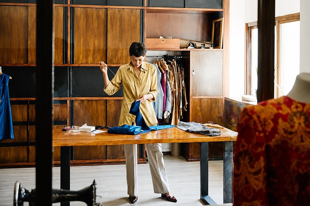 Wide editorial shot of an atelier or small production space. A tailor working by hand in natural light. The image that communicates something real is being made.