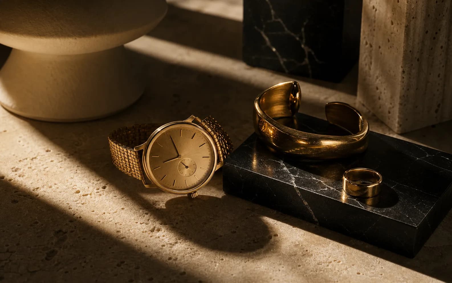 A refined gold-toned still life of watch and jewelry forms on warm stone.