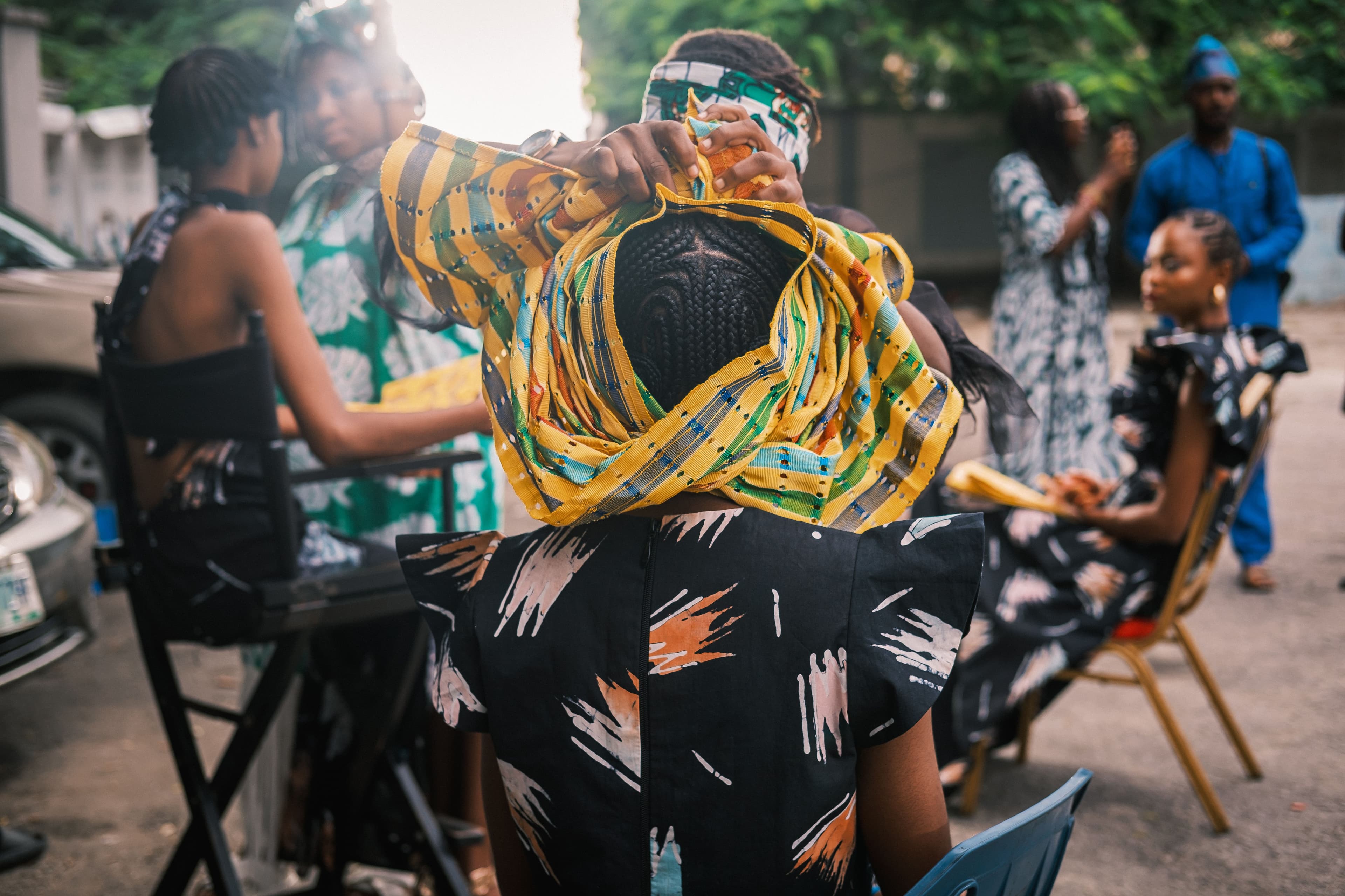 Model seen from behind in a black dress marked with white brushstrokes, a gold and green striped gele being tied above her on a Lagos street.